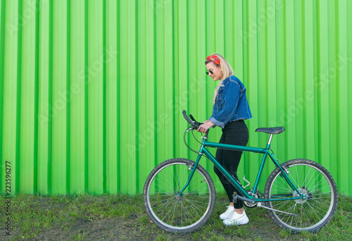 Portrait of a stylish woman in a denim jacket and sunglasses, pulling a bike on the background of a green wall. Copyspace