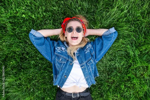 Emotional stylish woman in bright clothes lays on the grass, looks from above, looks into the camera and smiles. Happy emotional girl in stylish clothes lays on the lawn. Top view © bodnarphoto