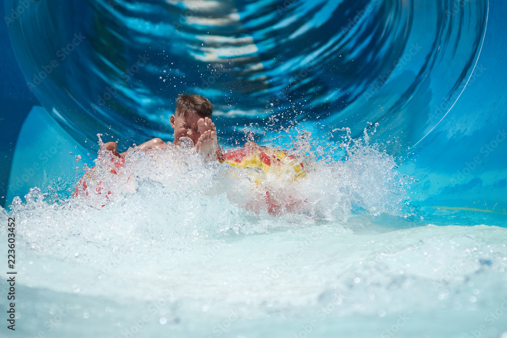 Caucasian boy gliding down slide in aqua park. Water splashes are all over.