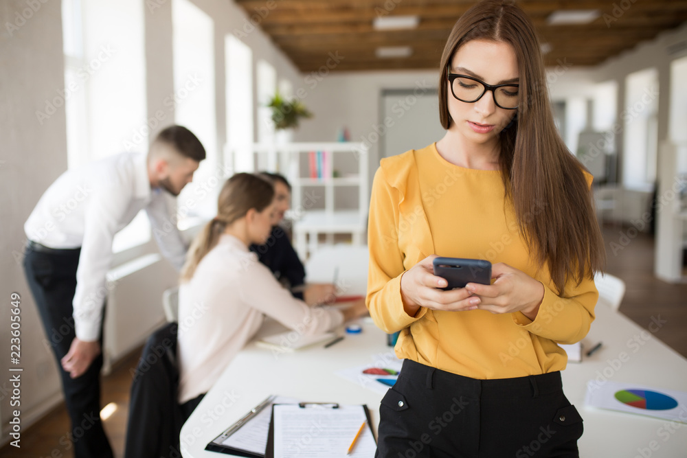 Beautiful business woman in eyeglasses using cellphone in office with colleagues on background Beautiful business woman in eyeglasses using cellphone in office with colleagues on background