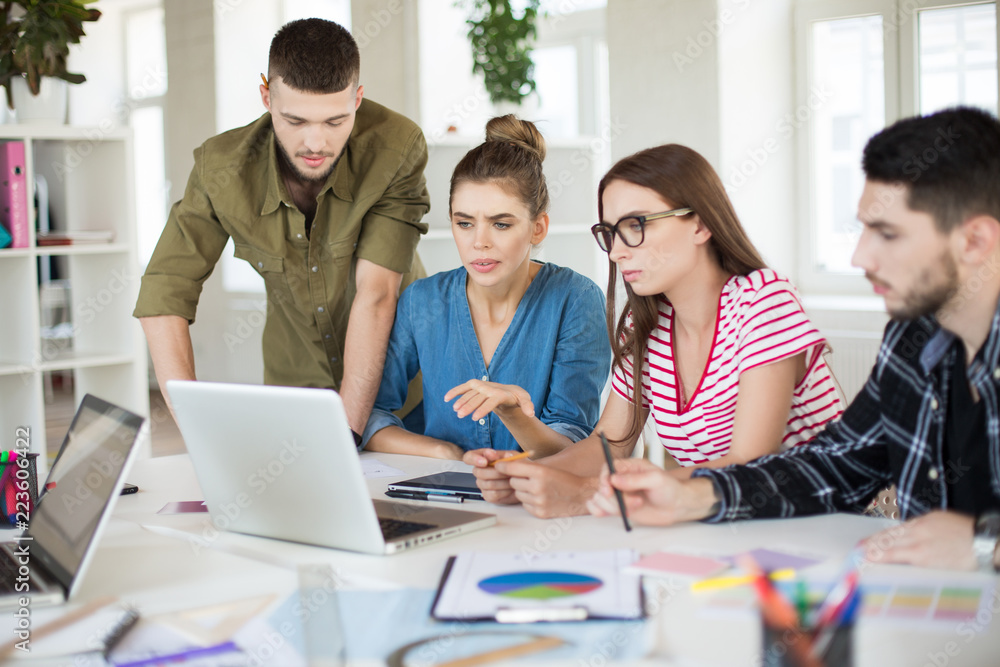 Group of young business people thoughtfully using laptop together. Pensive men and women spending time at work in office Group of young business people thoughtfully using laptop together. Pensive men and women spending time at work in office