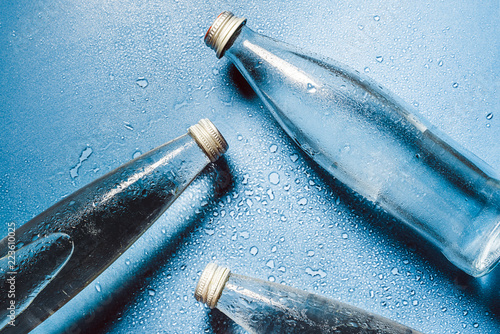 A glass bottle of water on blue background splashes drops of water on top. Quench thirst. Background of water drops