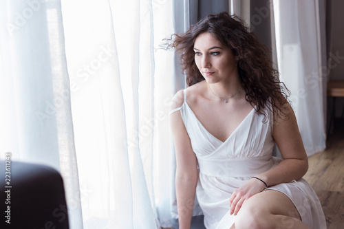 Young woman with curly hair sitting on the floor by the window, relaxing in her hotel room.