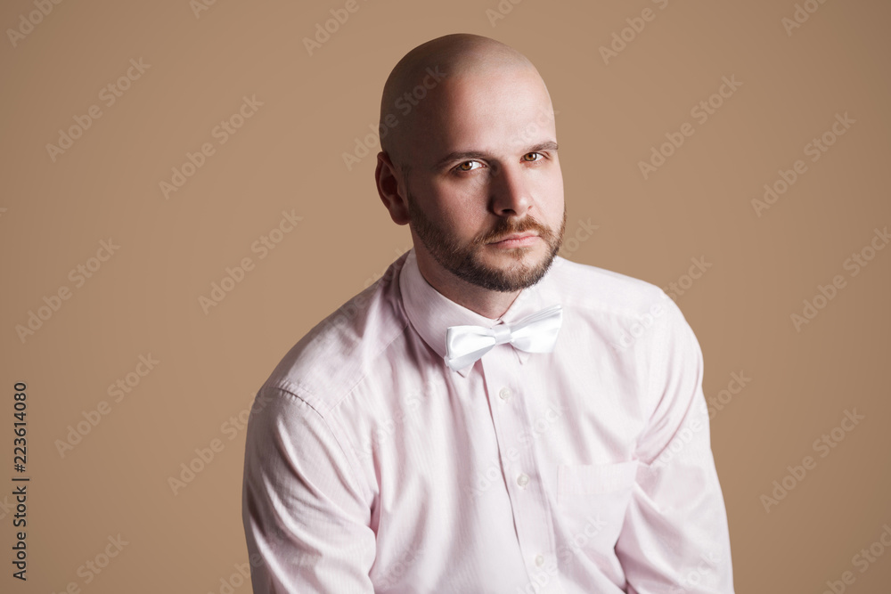 Portrait of handsome bearded bald man in light pink shirt and white bow, sitting on chair and looking at camera with serious face. indoor studio shot, isolated on brown background. Portrait of handsome bearded bald man in light pink shirt and white bow, sitting on chair and looking at camera with serious face. indoor studio shot, isolated on brown background.