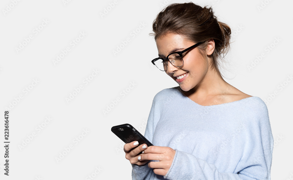Portrait of pretty joyful woman in stylish glasses posing at studio. Cute brunette holding modern cellphone in hands and looking at display with gladness. Isolated on grey background