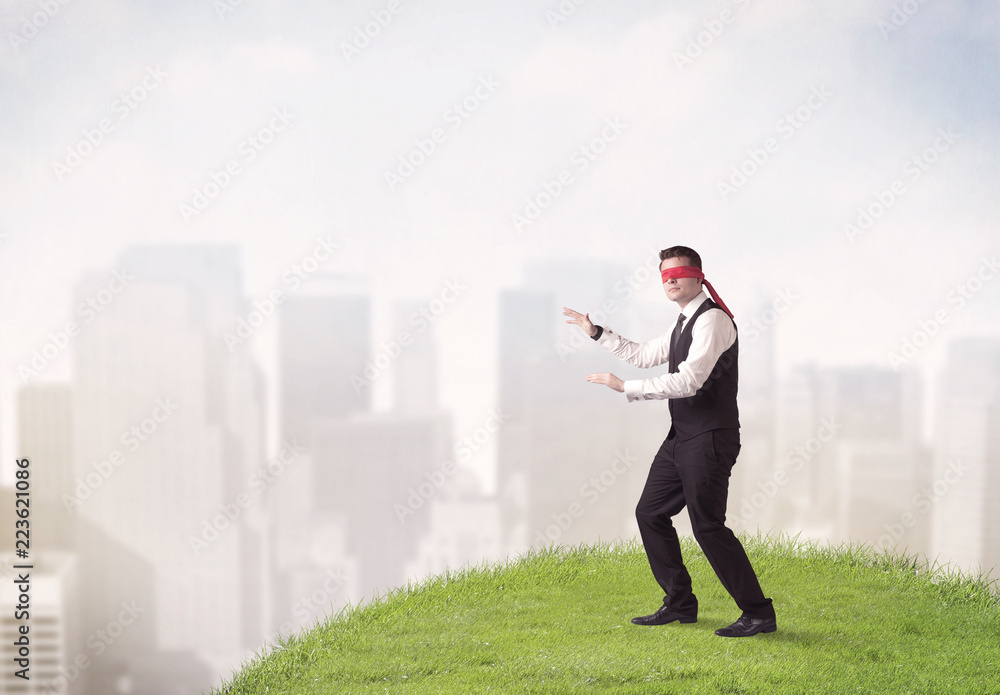 Young blindfolded businessman steps on a a patch of grass with a city in the background Young blindfolded businessman steps on a a patch of grass with a city in the background
