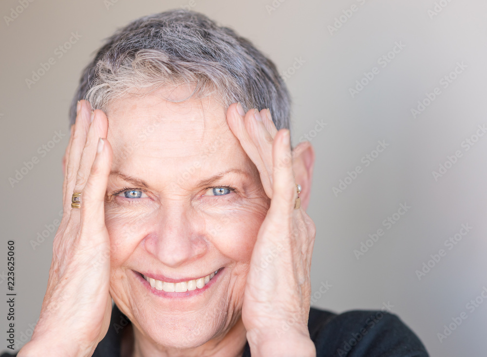 Close up portrait of beautiful older woman with short grey hair smiling with hands around face against neutral background (selective focus) Close up portrait of beautiful older woman with short grey hair smiling with hands around face against neutral background (selective focus)