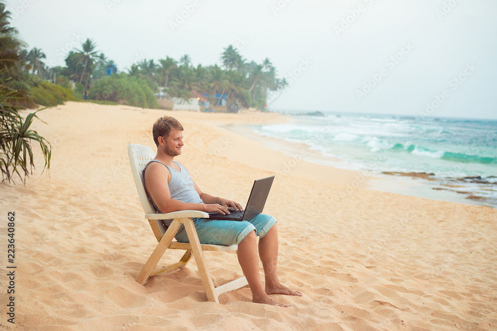 A young man with a laptop working on the beach. Remote work, freelance, Wi-Fi, technology, travel and leisure. Businessman with laptop on the beach, work on the beach during summer holidays .
