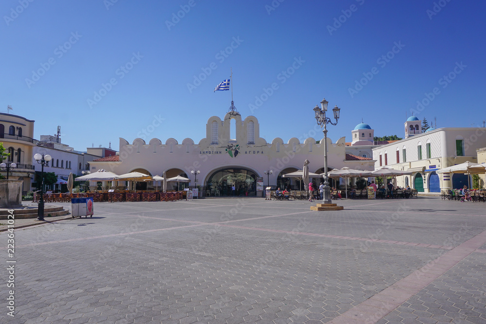 Fototapeta premium Kos, Greece: Cafes and shops lie around the edges of Eleftherias Square, with an indoor market hall at the center, in Kos Town.