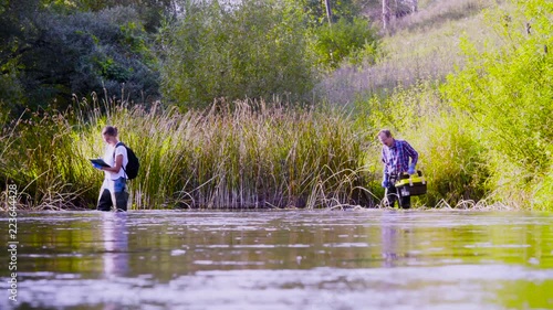 Crane shot. Two scientists ecologists in high rubber boots walking in the water of the forest river. Field work