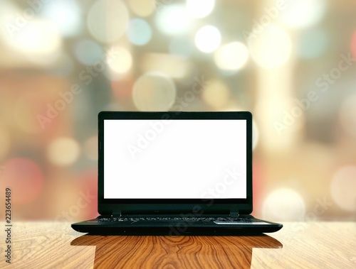 Black laptop and empty white screen on wooden glossy table with bokeh and blurred lighting in living room background on work space and technology concept 