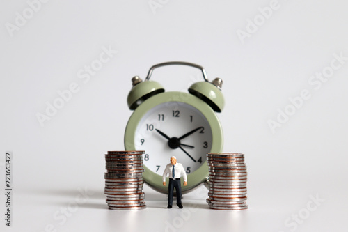 Miniature old man standing with a pile of coins in front of the alarm clock.