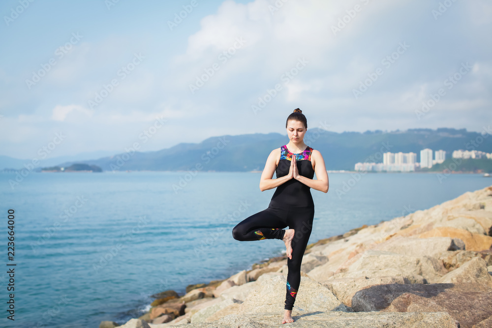 Beautiful young woman in sports jumpsuit doing yoga exercise outdoors with sea views