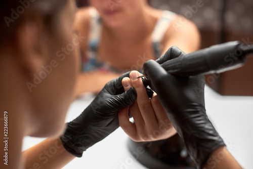Process of manicure making in beauty salon. Close-up of professional master manicurist hands in black gloves working on client fingernails, drilling and removing cuticle with electric file.