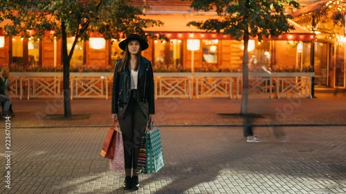 Time-lapse of young woman shopaholic standing outdoors in the street with shopping bags and looking at camera while flow of people is moving around her.