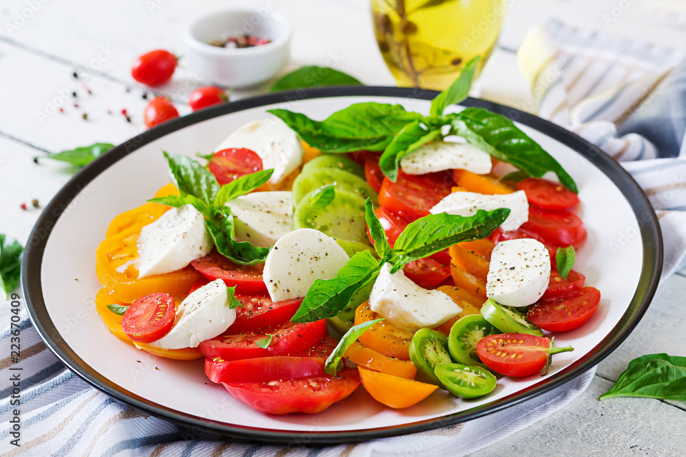 Mozzarella cheese, tomatoes and basil herb leaves in plate on the white wooden table. Caprese salad. Italian food. Mozzarella cheese, tomatoes and basil herb leaves in plate on the white wooden table. Caprese salad. Italian food.