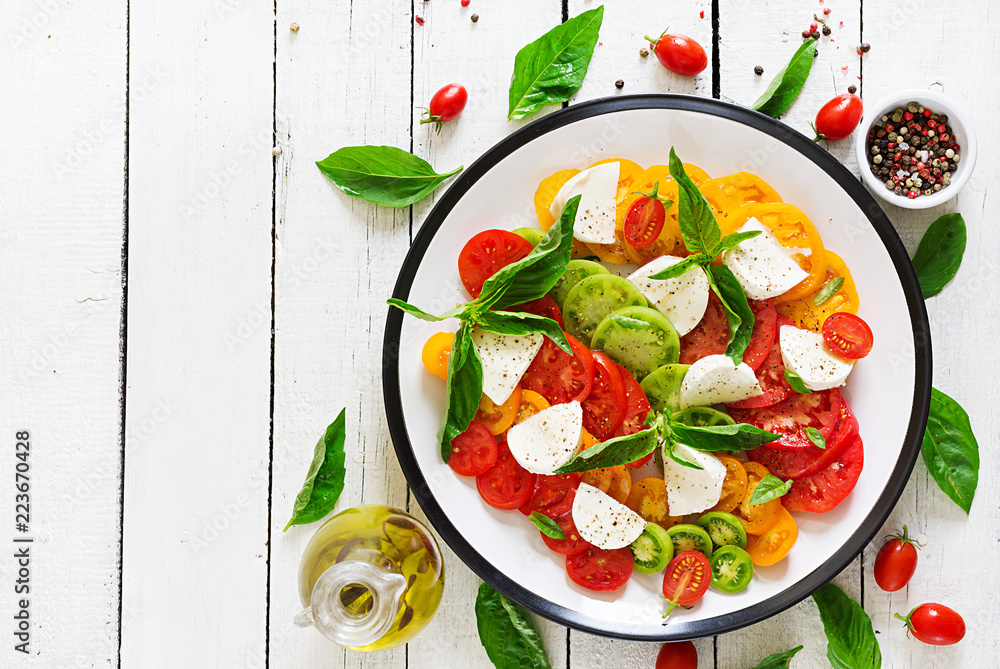Mozzarella cheese, tomatoes and basil herb leaves in plate on the white wooden table. Caprese salad. Italian food. Top view Mozzarella cheese, tomatoes and basil herb leaves in plate on the white wooden table. Caprese salad. Italian food. Top view
