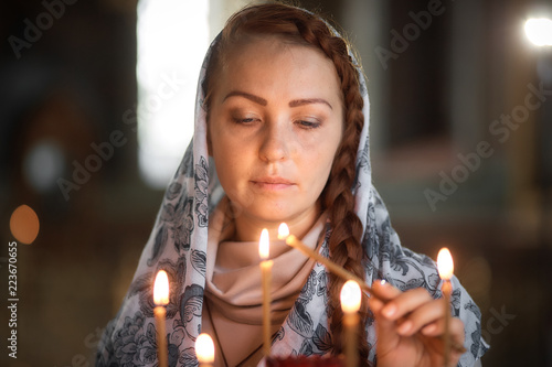 Russian beautiful Caucasian woman with red hair and a scarf on her head is in the Orthodox Church, lights a candle and prays in front of the icon
