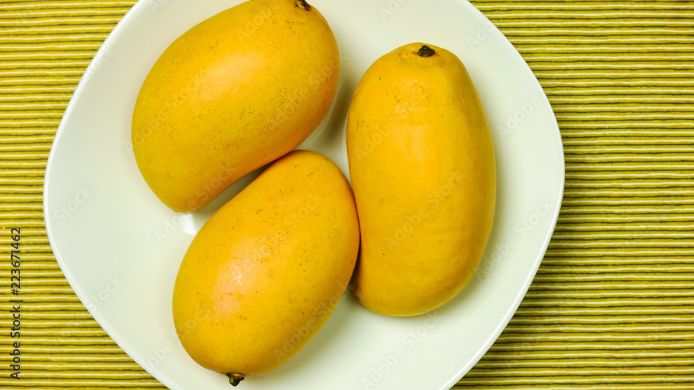 Three bright, yellow mangoes on a white plate on a striped background