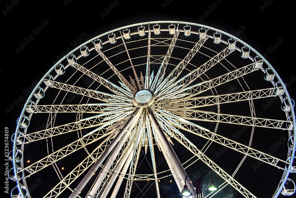 A giant ferris wheel at night lit up with white and yellow lights running all over the structure. A lamp post is visible in the foreground and the view is looking up from the base.