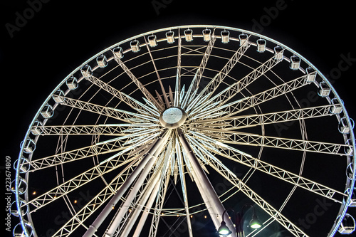 Obraz na płótnie A giant ferris wheel at night lit up with white and yellow lights running all over the structure. A lamp post is visible in the foreground and the view is looking up from the base. w salonie