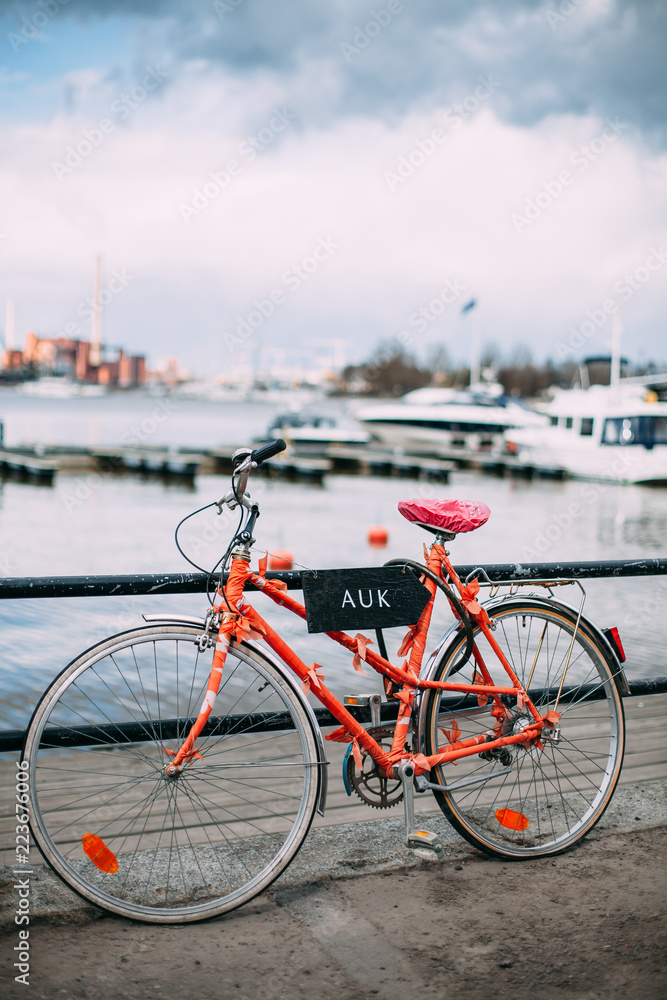 A red bicycle stands at the fence in the city port. In the background there are yachts, boats, ships in the bay. Cloudy sky over the bay. The sea in the city