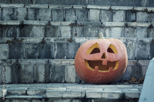 A large orange pumpkin with a carved face. The pumpkin stands on the steps in the sinister twilight.