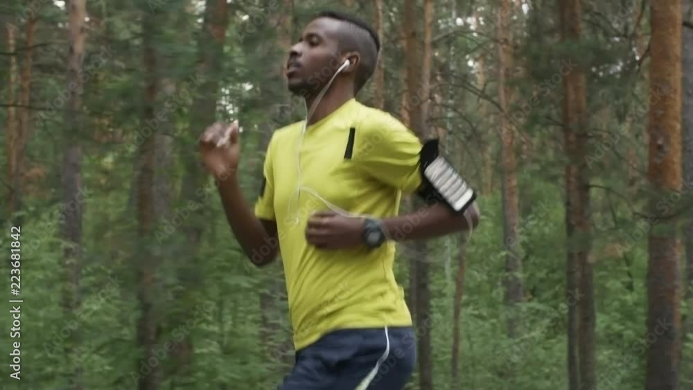 Tracking shot with tilt down of determined black man listening to music in earphones and running on pavement in park in morning Tracking shot with tilt down of determined black man listening to music in earphones and running on pavement in park in morning