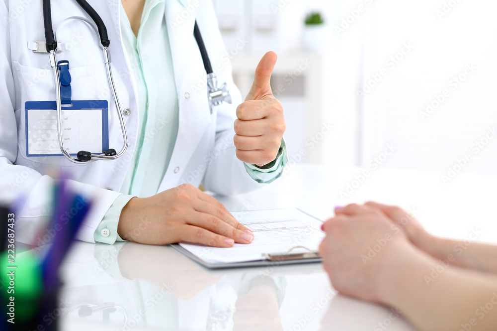 Doctor showing Ok sign with thumb up to patient while sitting at the desk in hospital office, close-up of human hands. Medicine and health care concept Doctor showing Ok sign with thumb up to patient while sitting at the desk in hospital office, close-up of human hands. Medicine and health care concept