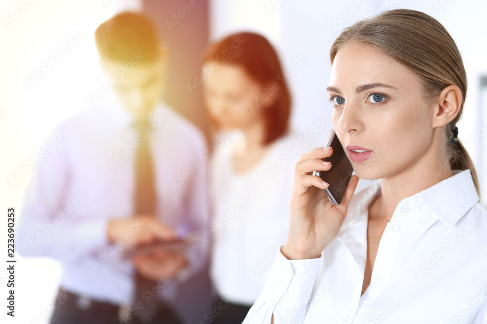 Serious blonde businesswoman talking by phone in the background of her colleagues in office. Business concept Serious blonde businesswoman talking by phone in the background of her colleagues in office. Business concept