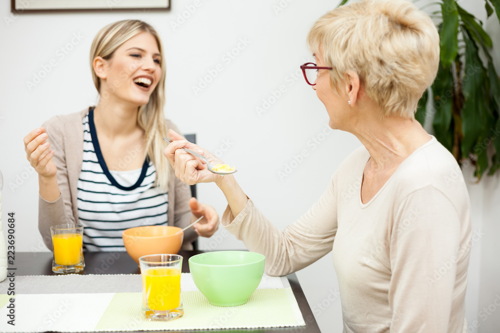 Senior mother and daughter talking and eating healthy cereal breakfast in brightly lit dining room. Focus on mother. Happy family moments at home Senior mother and daughter talking and eating healthy cereal breakfast in brightly lit dining room. Focus on mother. Happy family moments at home