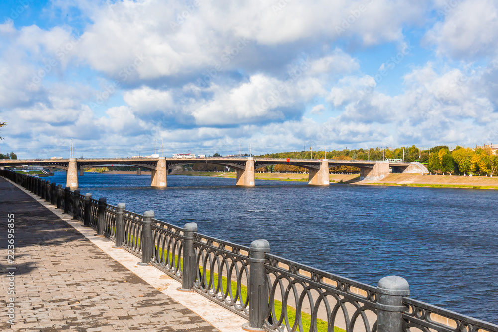 The new Volga bridge and city quay in Tver, Russia. Picturesque river landscape. Clouds in the blue sky. Summer or autumn Sunny day. Metal fence.