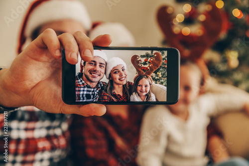 Christmas. Family. Technology. View on the smartphone. Dad, mom and daughter in Santa hats looking at camera and smiling while doing selfie