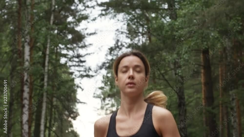 Dolly shot of determined young woman wearing sports top running in forest in morning Dolly shot of determined young woman wearing sports top running in forest in morning