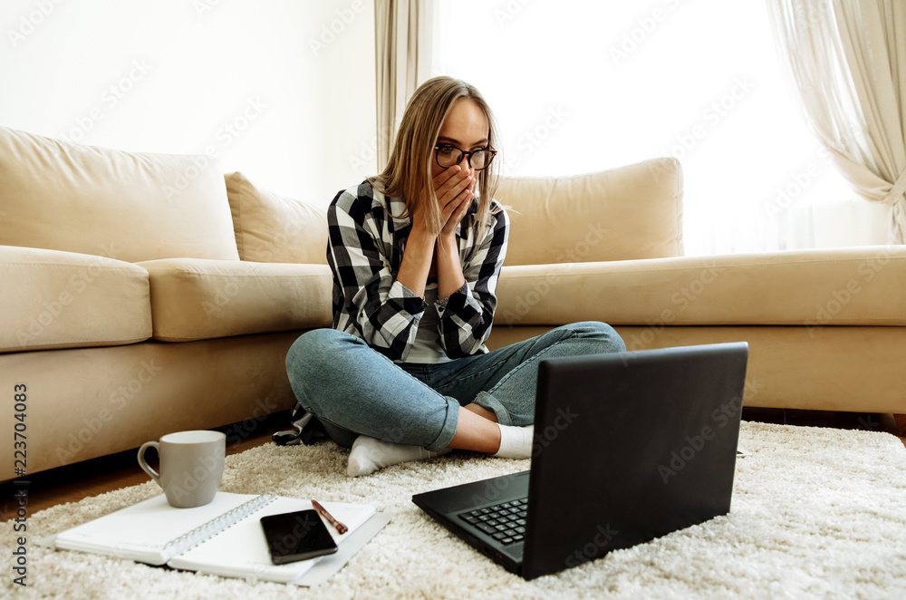 Shock. Education. Technology. Home. Girl in casual clothes and eyeglasses is covering her mouth and looking at a laptop with widely open eyes; at home