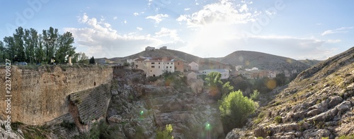 A landscape at the sunset of the rural town of Almonacid de la Cuba, in Spain, with houses on a small valley with the Aguavivas river and waterfall. An ancient Roman bridge is crossing the river.