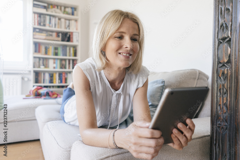 Smiling mature woman lying on couch at home using tablet Smiling mature woman lying on couch at home using tablet