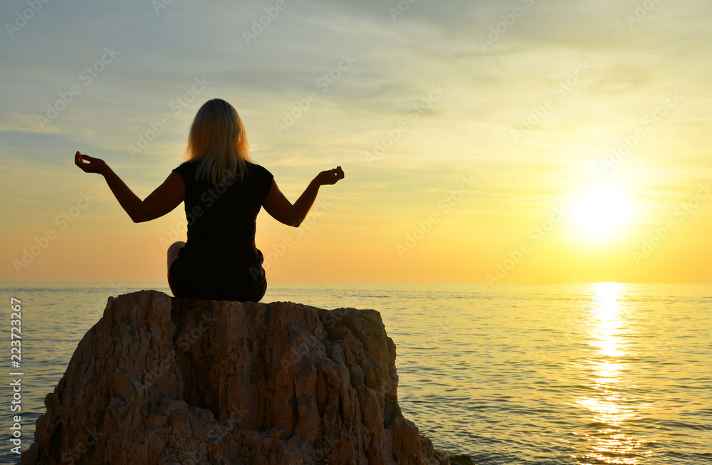 Silhouette of a girl practicing yoga on a cliff by the sea at sunset.
