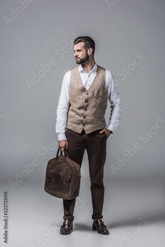 Fototapeta stylish elegant man in waistcoat posing with leather bag on grey