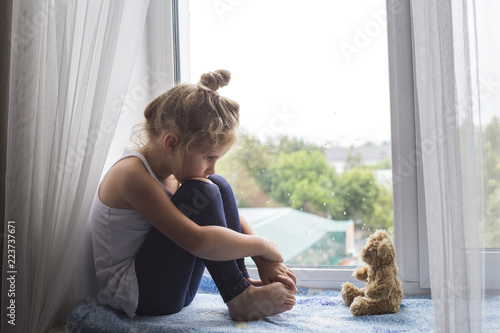 Sad litttle girl sits on a window sill and looks at a bear