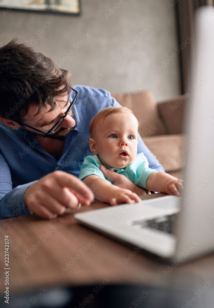 Family - Father using a laptop computer for work at home while looking after his baby son. Family - Father using a laptop computer for work at home while looking after his baby son.