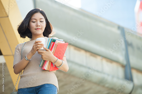 Beautiful Asian female college student holding her books and a cup of coffe standing outdoor, people education learning high school program smart teenager concept