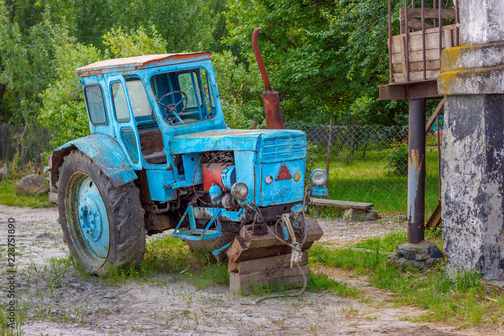 The abandoned rusty tractor without front wheels on the russian village street