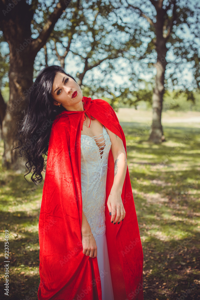 Halloween concept, glamorous costume detail. Young beautiful and mysterious woman in woods, in white Dress and Red Cloak. Little red hood or vampire story

 