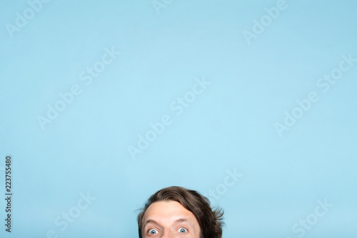 startled eccentric whimsical man with smiling eyes peeping out from the bottom of blue background. emotion and facial expression concept.