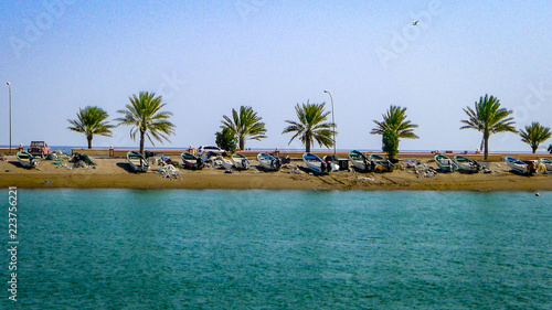 A row of palm trees and empty boats sit on a sand bank in front of blue waters at the fishing village of Quriyat in Oman photo