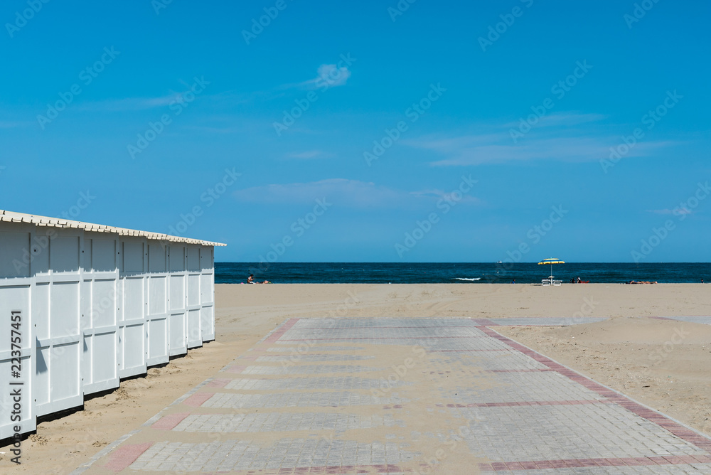 ein fast leerer Strand an der Adria mit weißen Badehütten, am Horizont ein blau-gelber Sonnenschirm