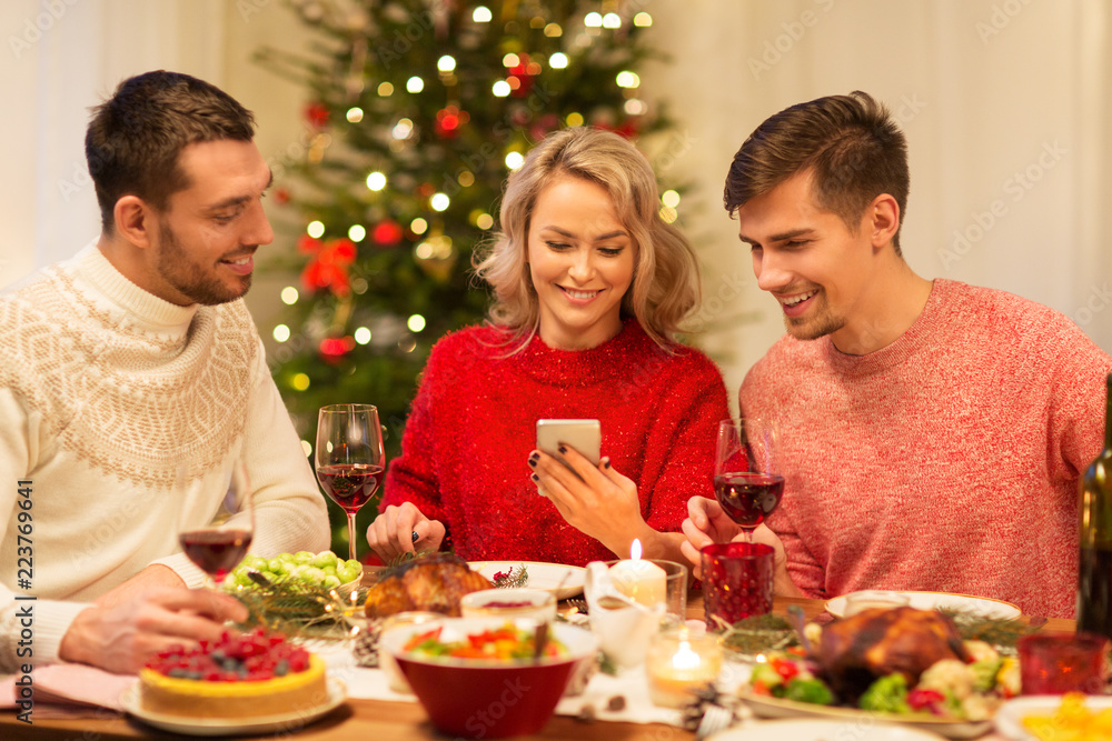 holidays and celebration concept - happy friends with smartphone having christmas dinner at home