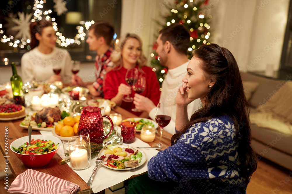 holidays, communication and celebration concept - happy young woman calling on smartphone and having christmas dinner with friends at home