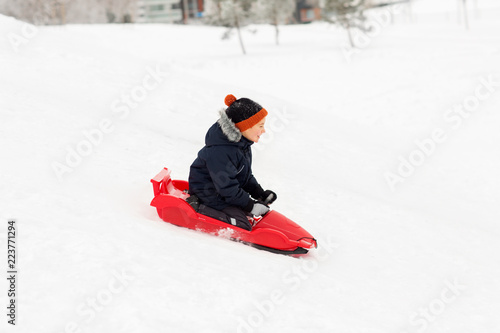 childhood, sledging and season concept - happy little boy sliding on sled down snow hill outdoors in winter